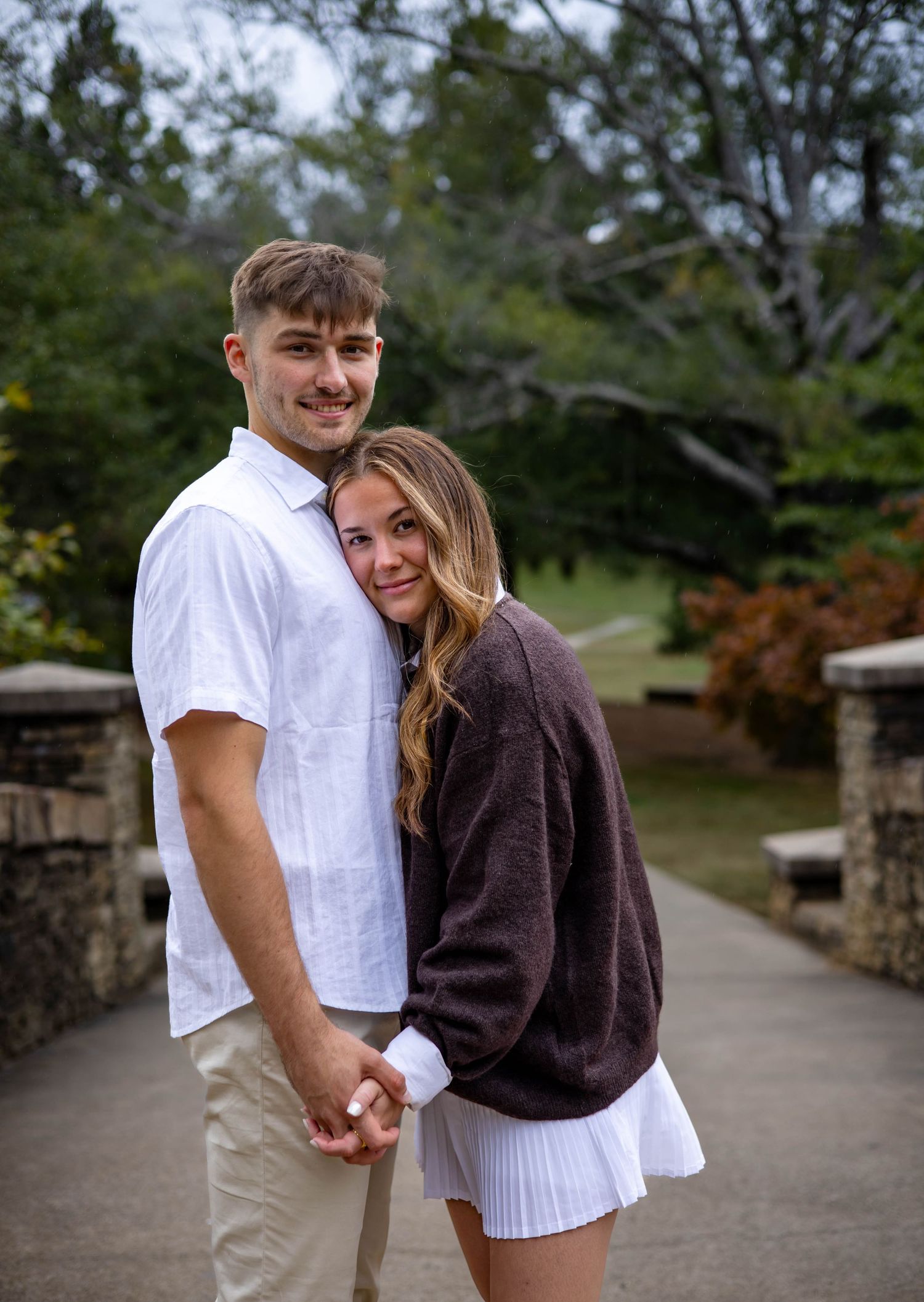 A couple embraces on a stone bridge pathway surrounded by lush greenery on an overcast day.