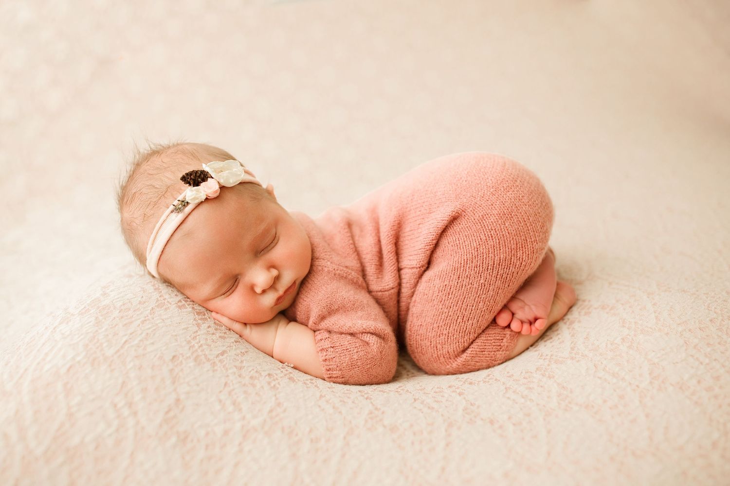 Newborn baby wearing pink wrap and delicate headband sleeps peacefully on cream colored blanket.