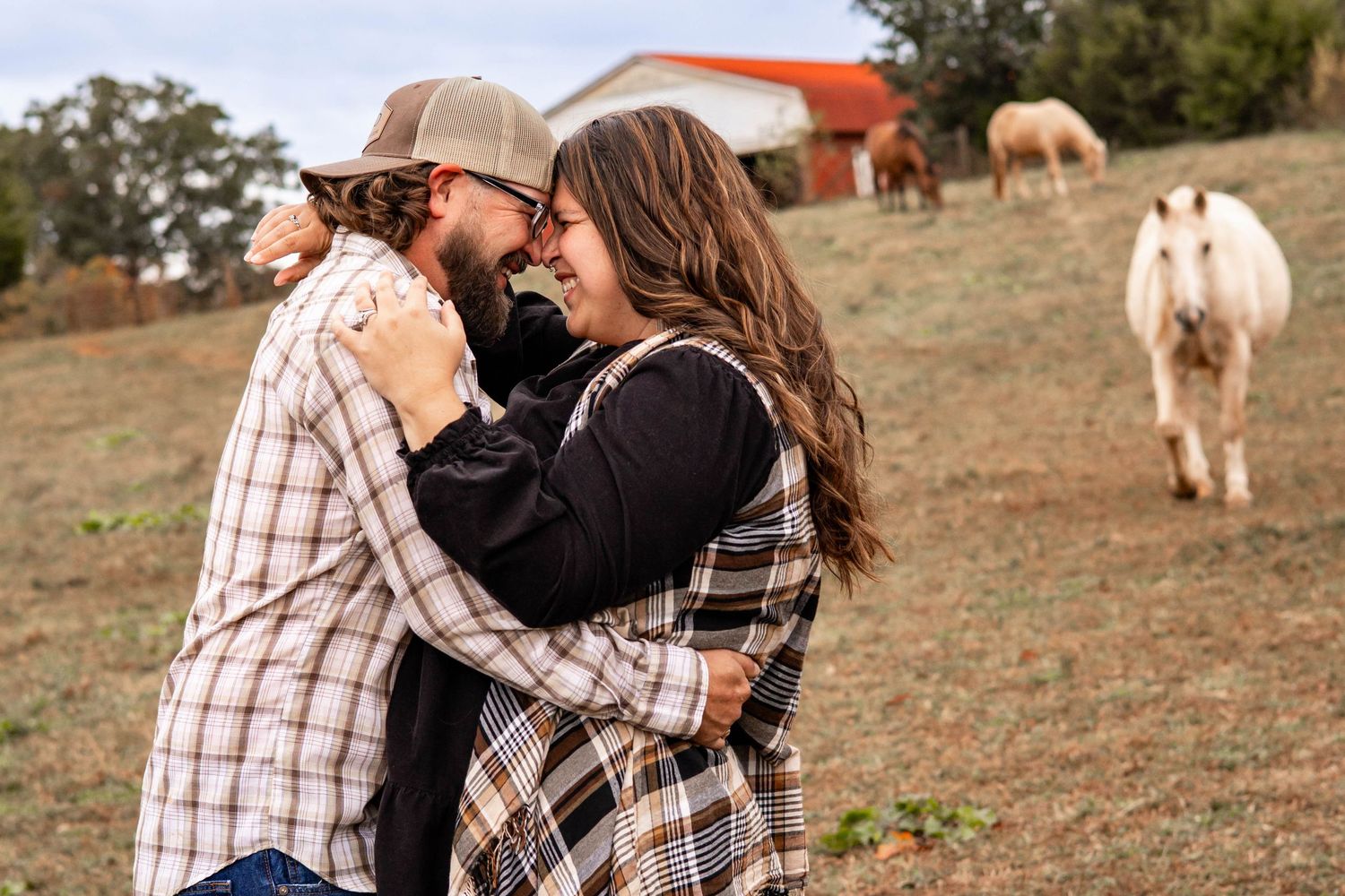 Happy couple embracing in a sunny field with horses grazing in the background during engagement photoshoot.