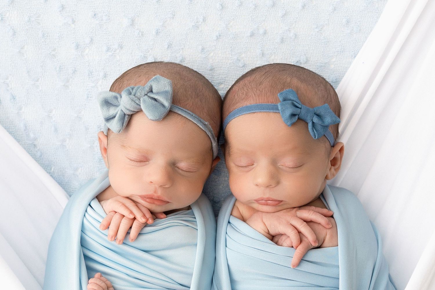 Two sleeping newborn babies wearing matching light blue outfits and headbands rest peacefully side by side.