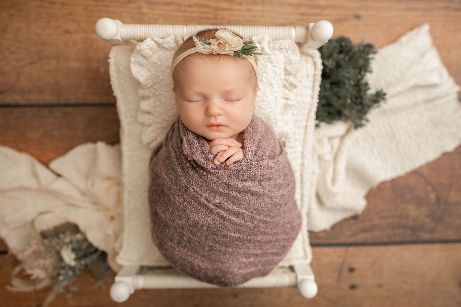 Newborn baby wrapped in mauve swaddle sleeping peacefully on white mini bed with floral headband.