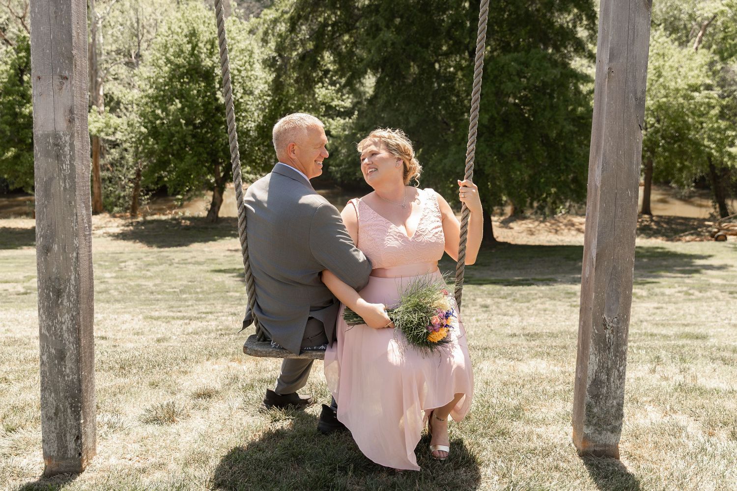 Older couple sitting on a wooden swing in a garden, woman in pink dress holding flowers, man in gray suit.