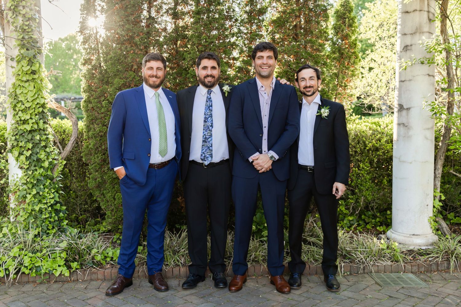 Four men in suits smiling together outdoors at a wedding, surrounded by lush green foliage and white columns.
