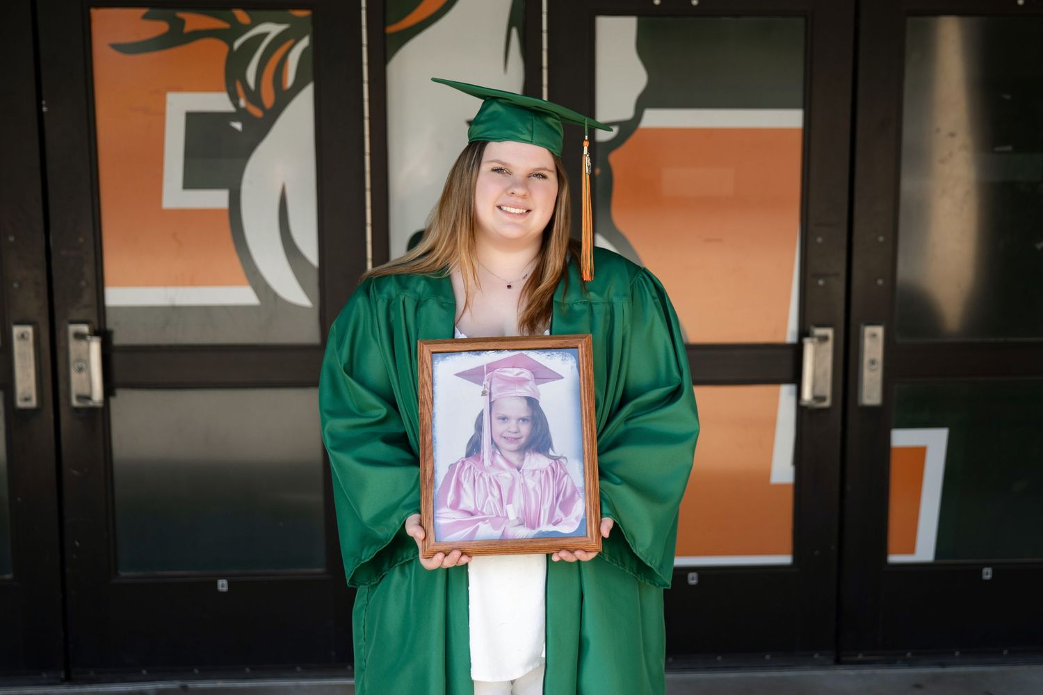 Graduate in green cap and gown holds framed portrait photo outside school with orange and black colors.