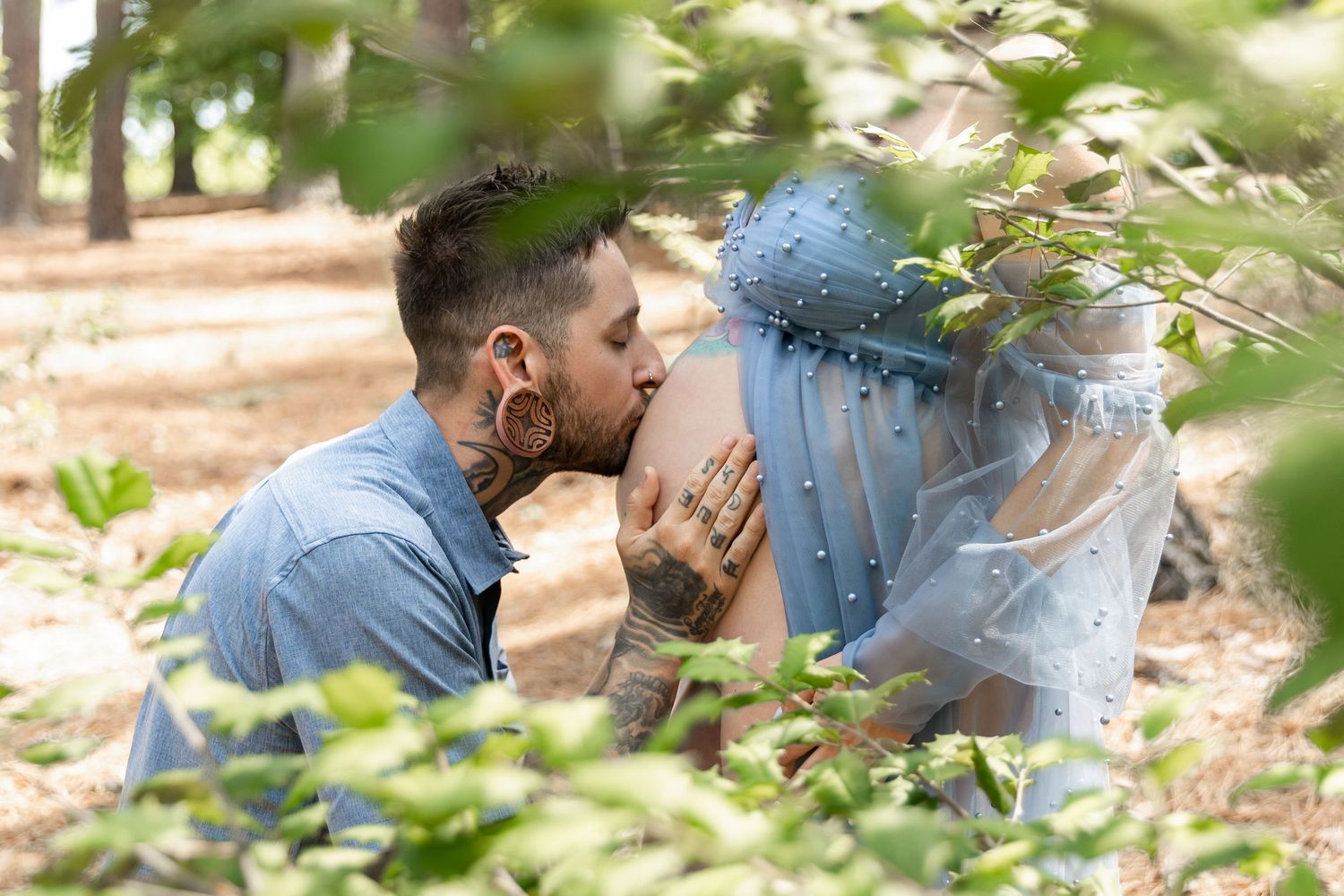 Tattooed man kissing pregnant woman's belly outdoors in a forest setting during a maternity photoshoot.