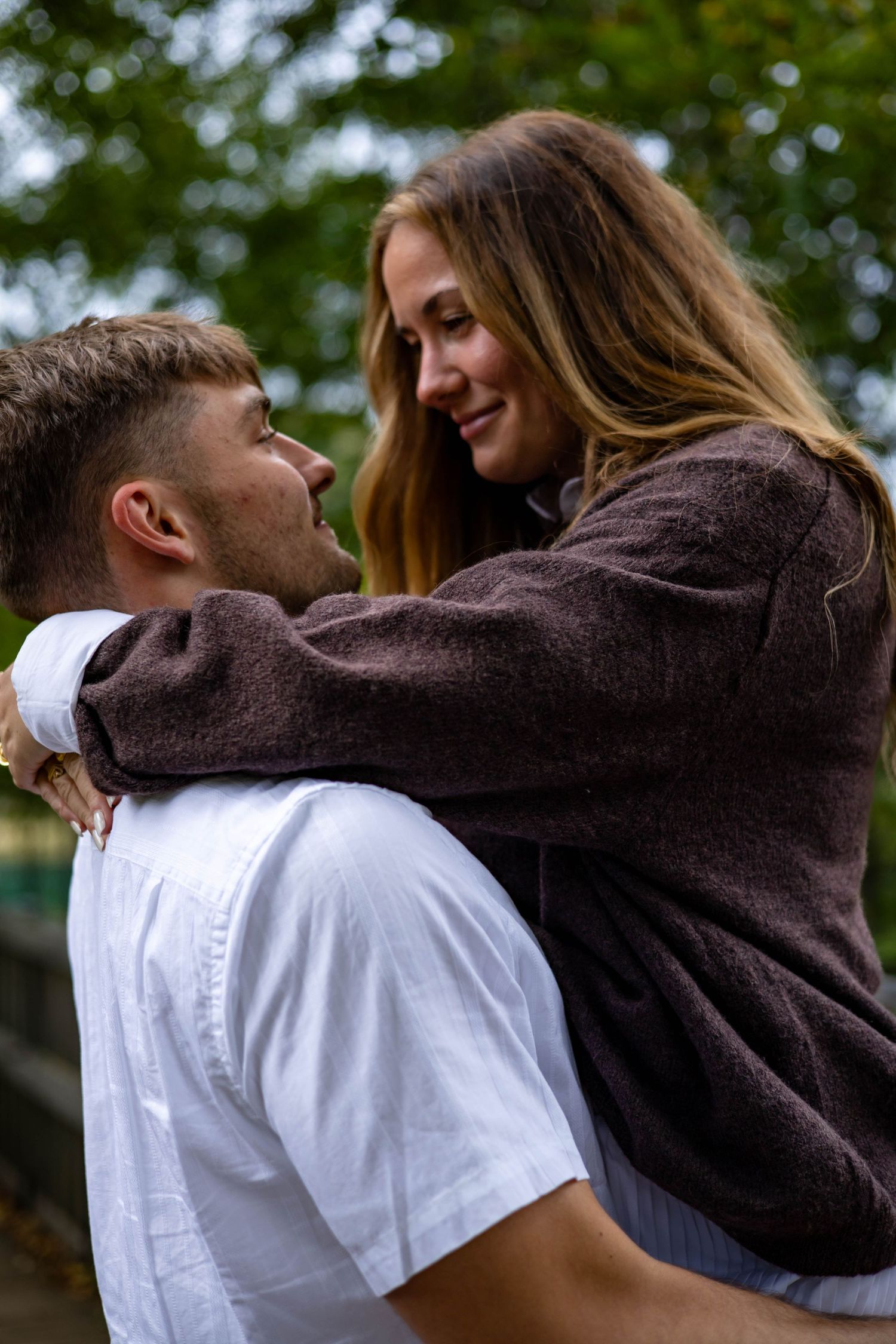 A couple shares a romantic moment outdoors, with one wearing a brown sweater embracing another in a white shirt.