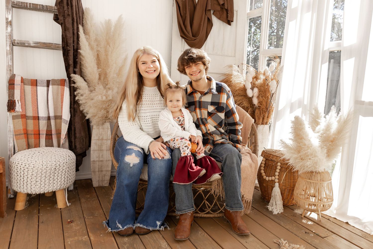 A woman, a teenage boy and a toddler sit together on a wooden bench in a bright boho studio with pampas grass and plaid blankets.