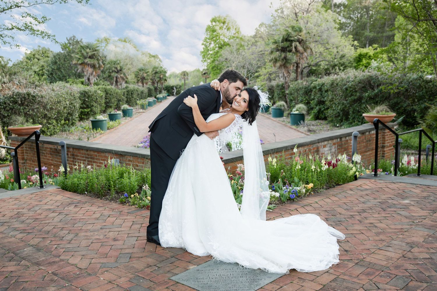 Bride and groom share a romantic dip kiss in a lush garden wedding venue with blooming flowers.