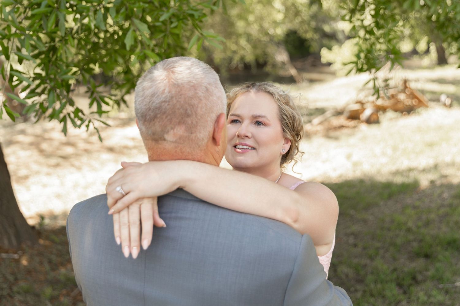 Bride in white dress smiling over groom's shoulder during outdoor wedding embrace under trees.