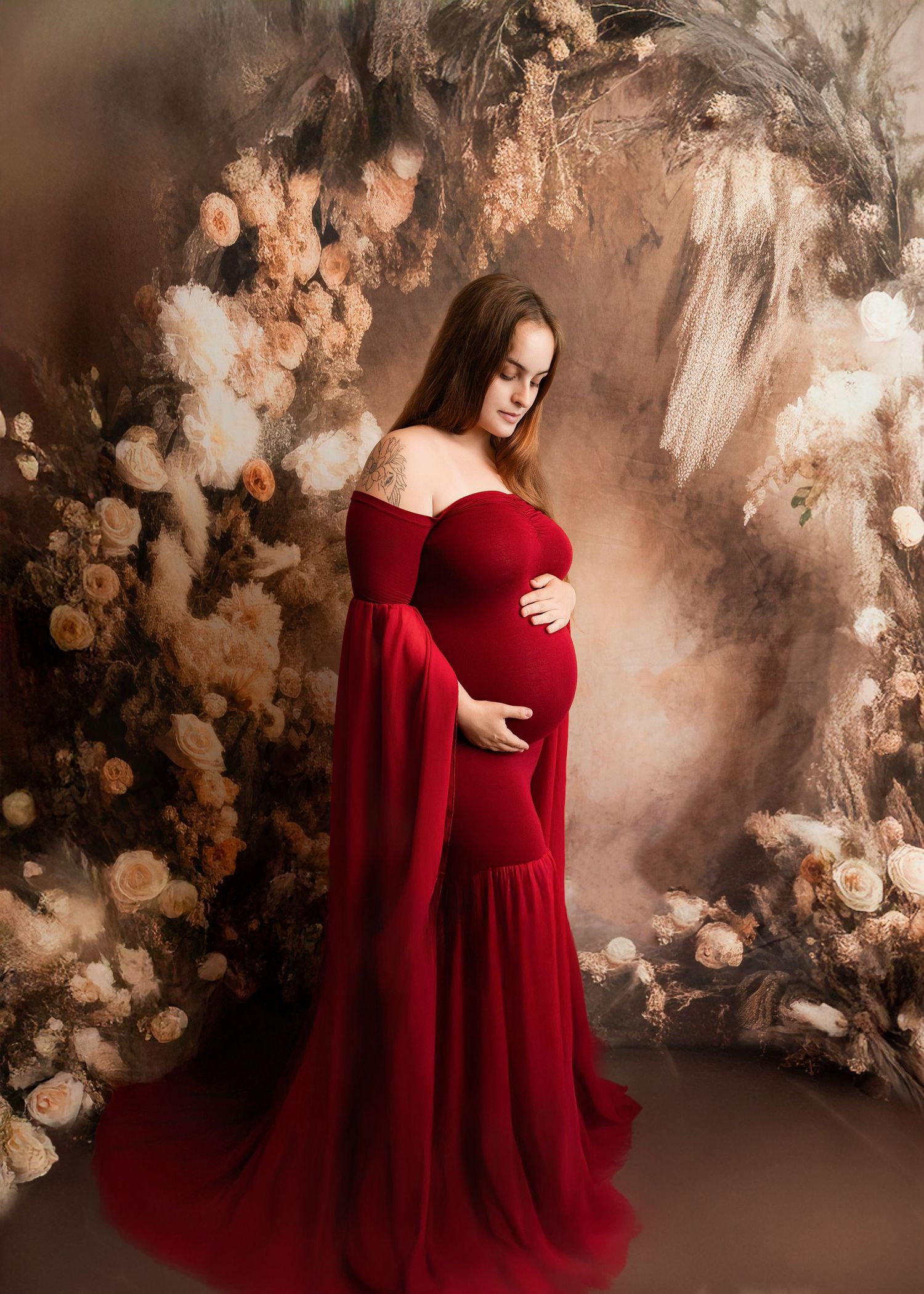 Pregnant woman in elegant red off-shoulder gown cradling baby bump, posed before a floral painted backdrop.