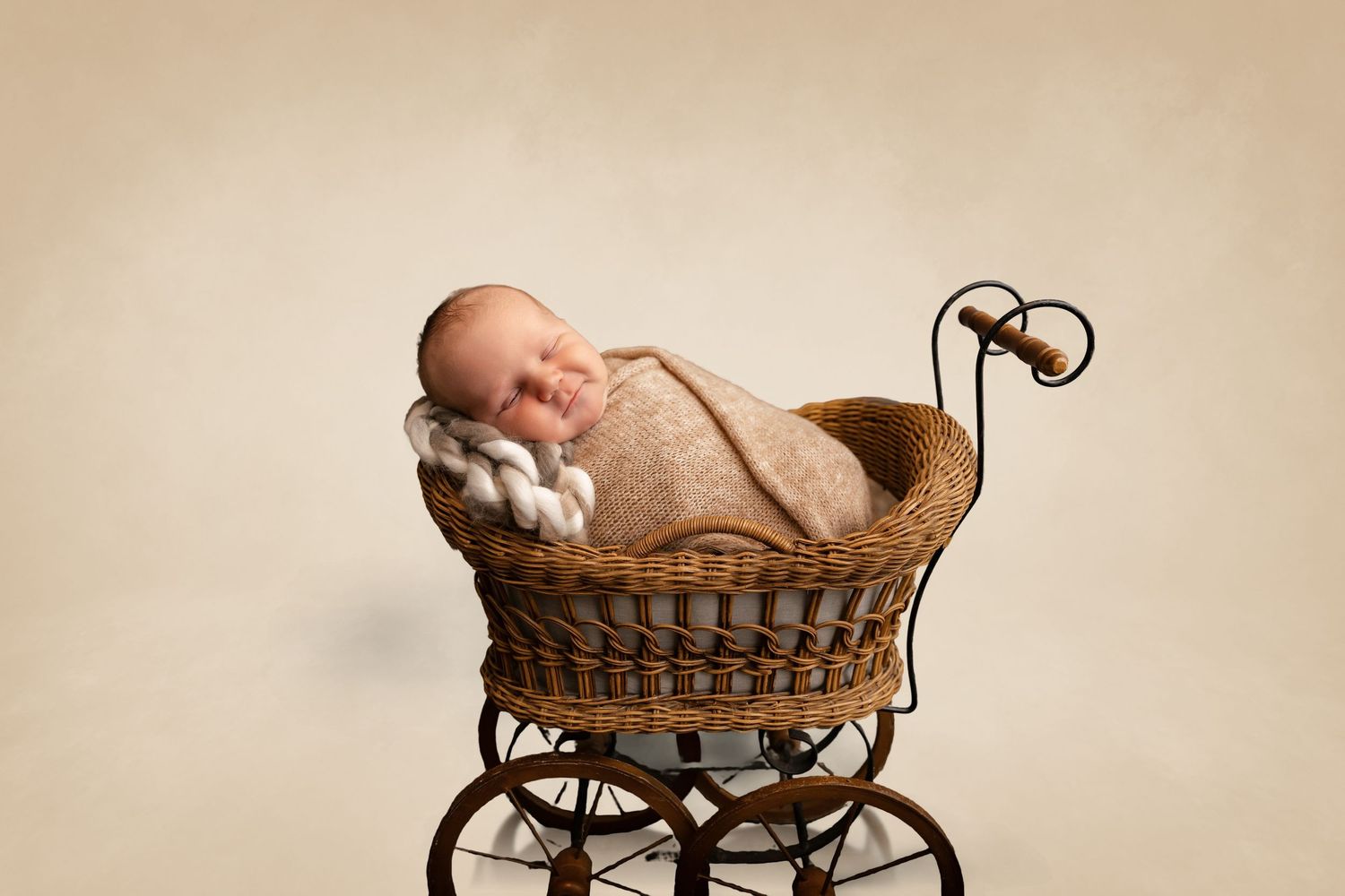 Newborn baby swaddled in beige wrap sleeping peacefully in a vintage wicker pram on a neutral background.