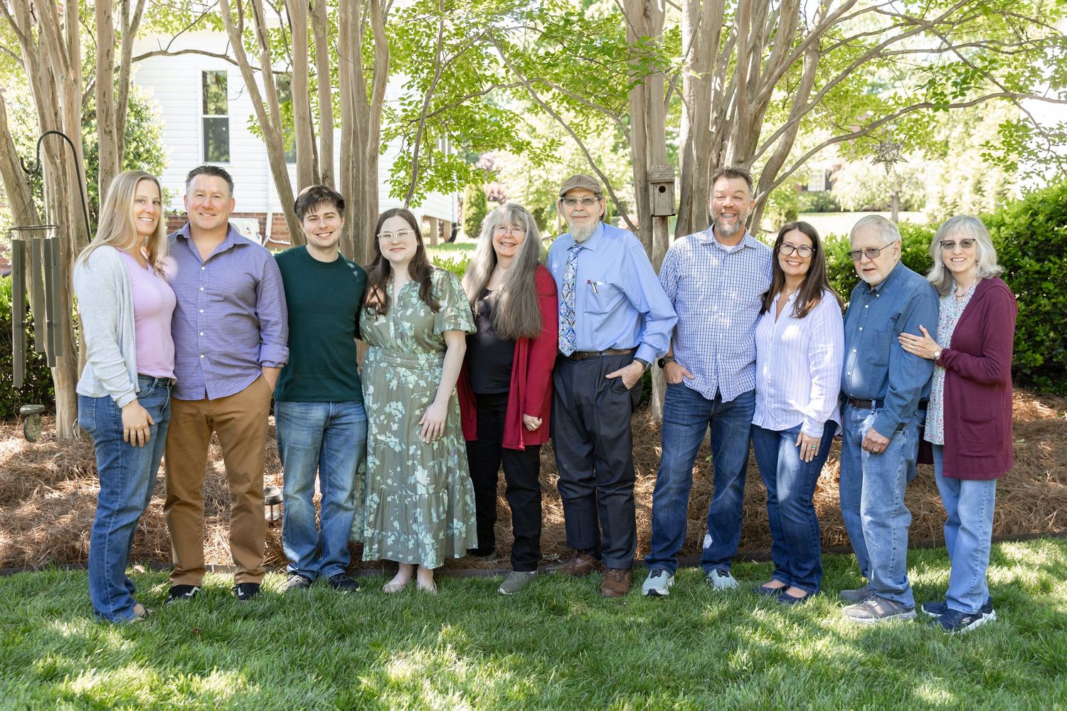 Group of ten adults smiling outdoors on green lawn surrounded by trees near a white building on a sunny day.