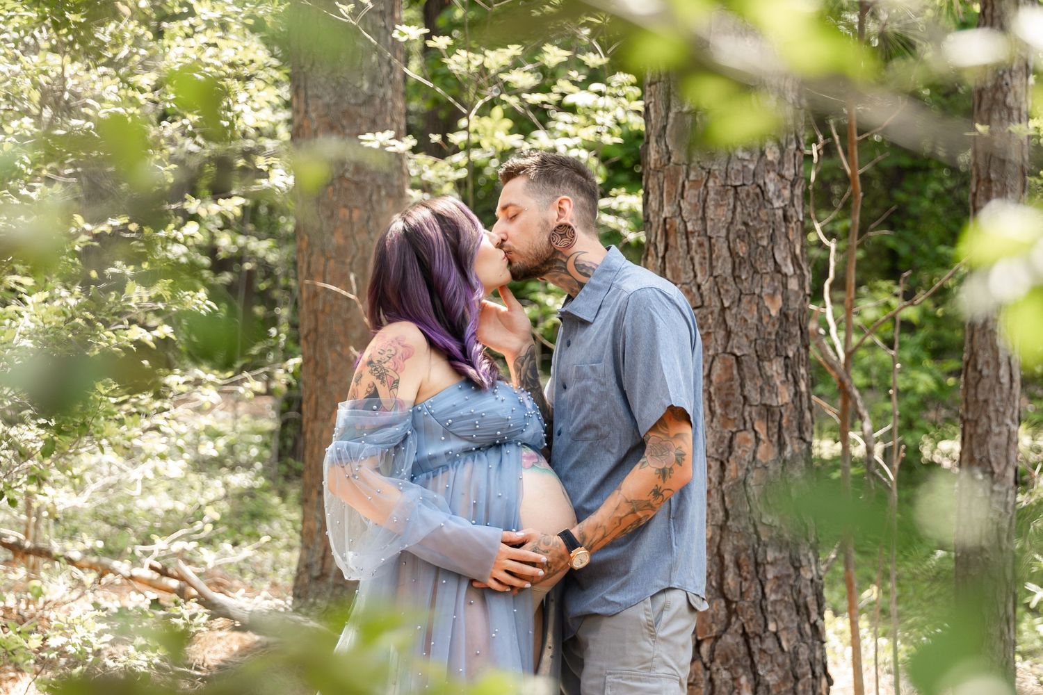 Pregnant couple kissing in a sunlit forest, woman with purple hair wearing a blue dress, man with tattoos embracing her baby bump.