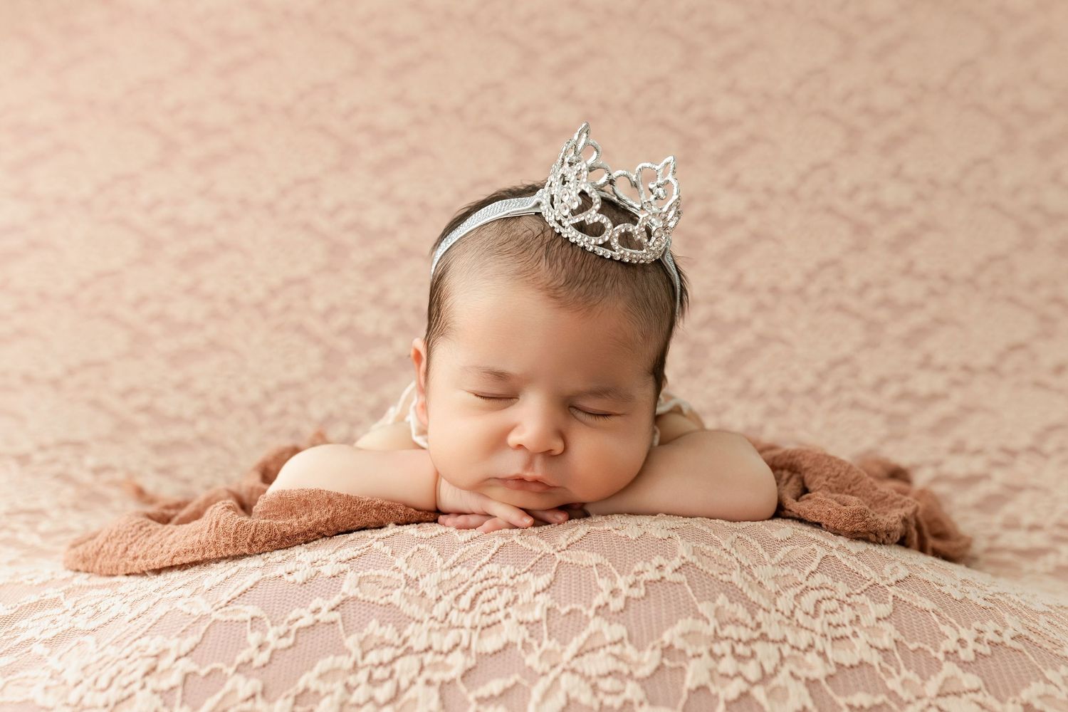 Newborn baby girl sleeping peacefully on lace fabric wearing a delicate silver tiara crown headband.