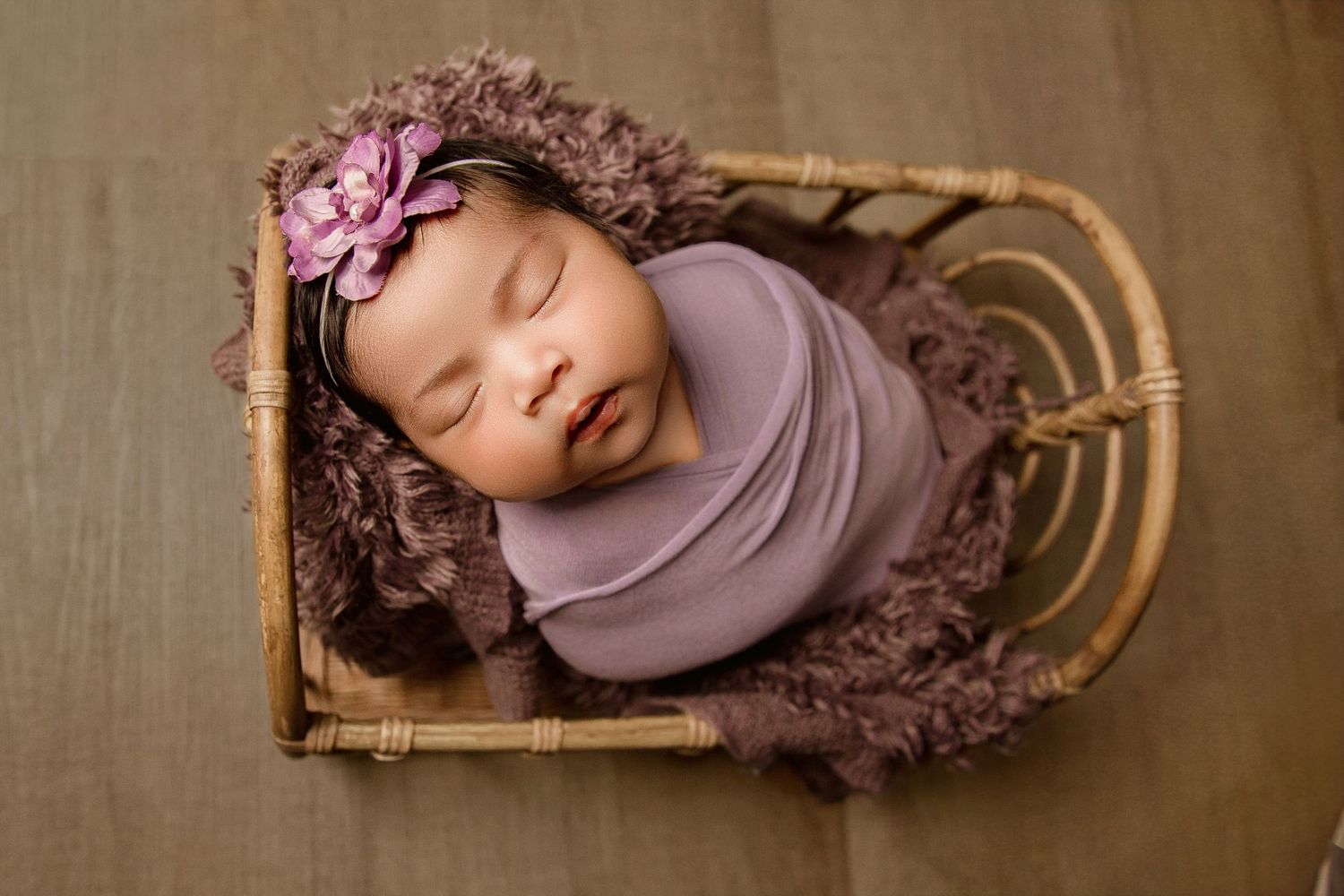 Sleeping newborn in brown basket with soft pink wrapping on textured backdrop.