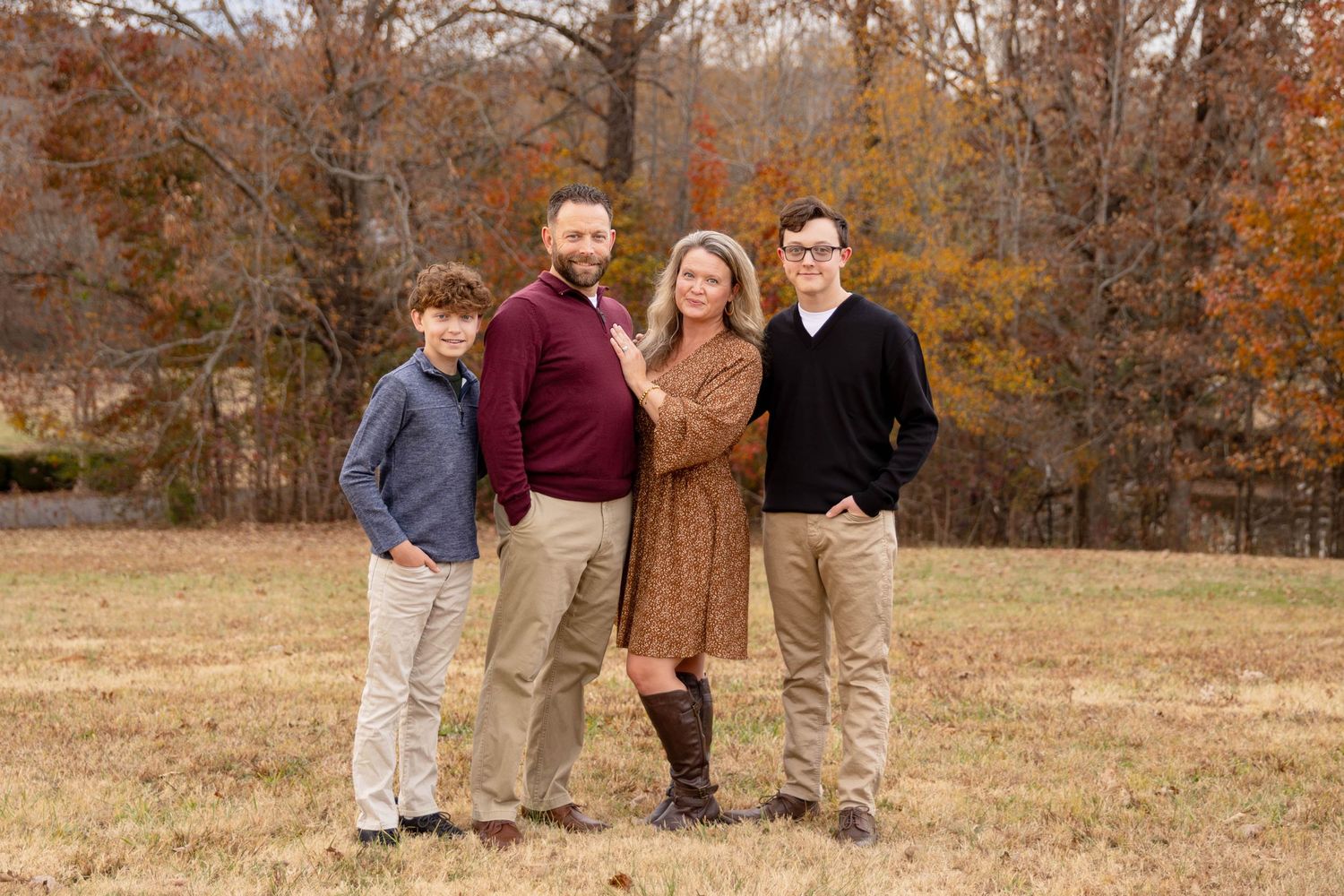 A family of four poses together in a fall field with orange and brown autumn trees in the background.