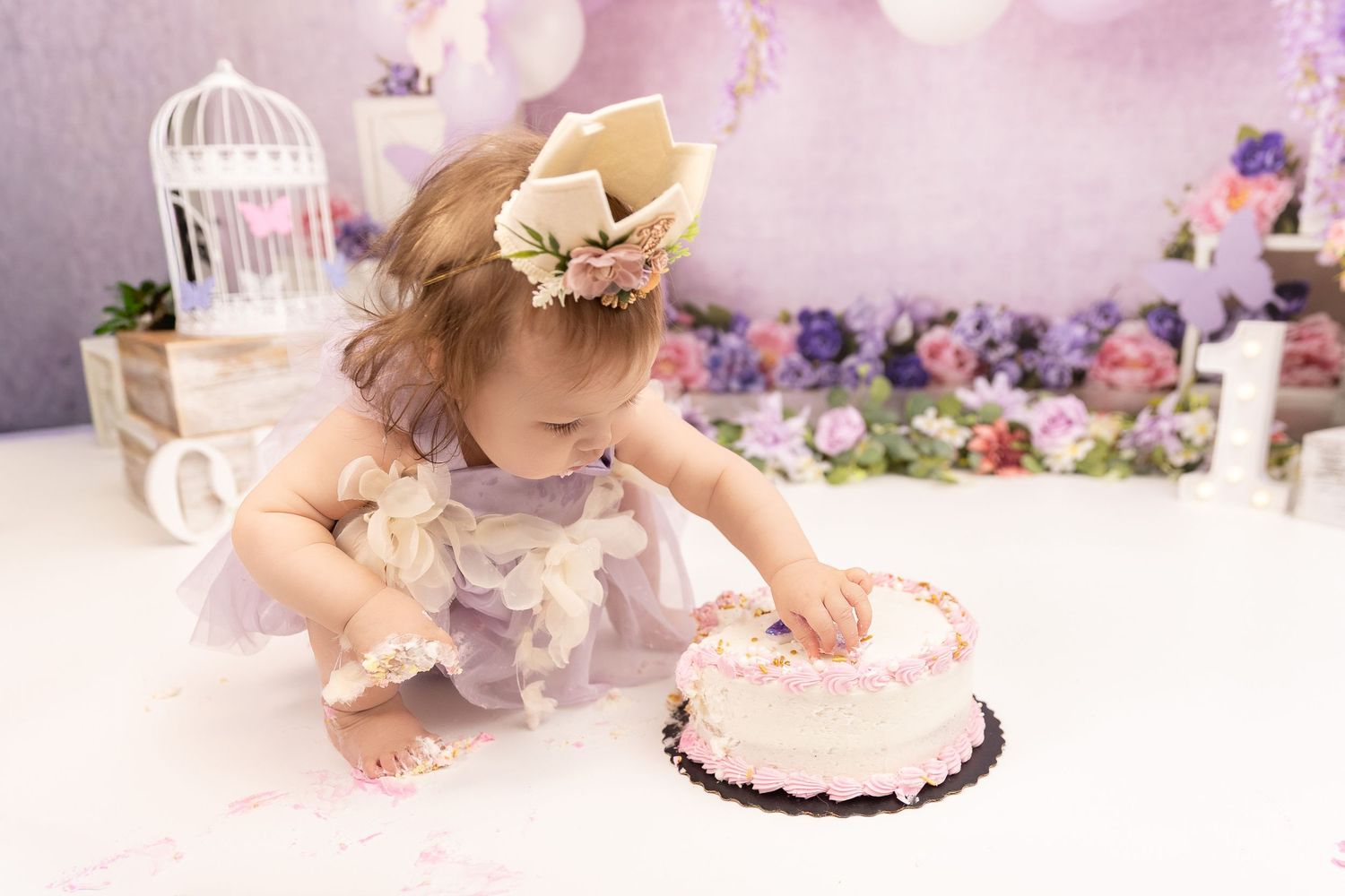 A baby in a vintage-style dress reaches for a pink birthday cake during a dreamy first birthday photo session.