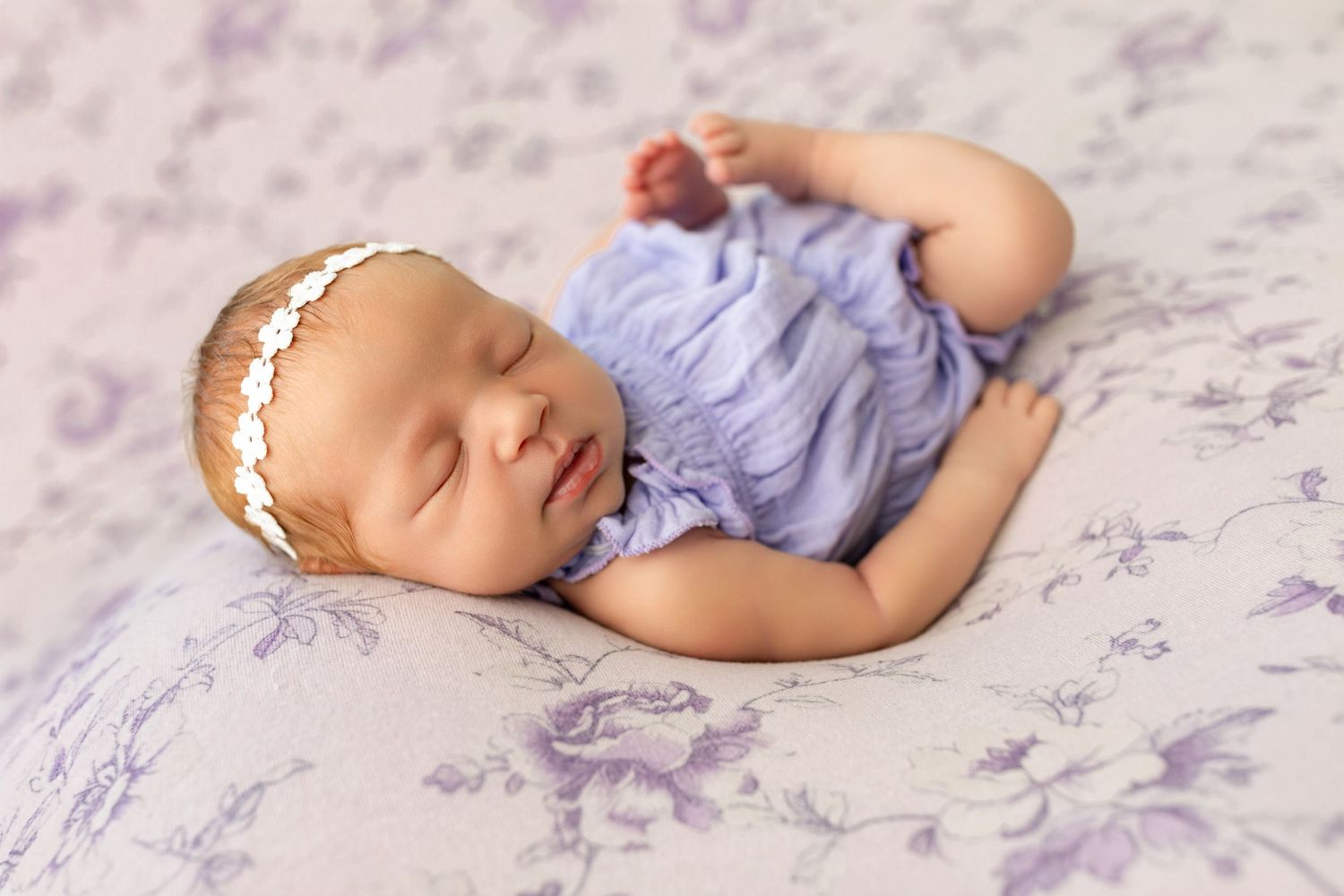 Sleeping newborn baby girl in lavender romper and floral headband on purple floral blanket.