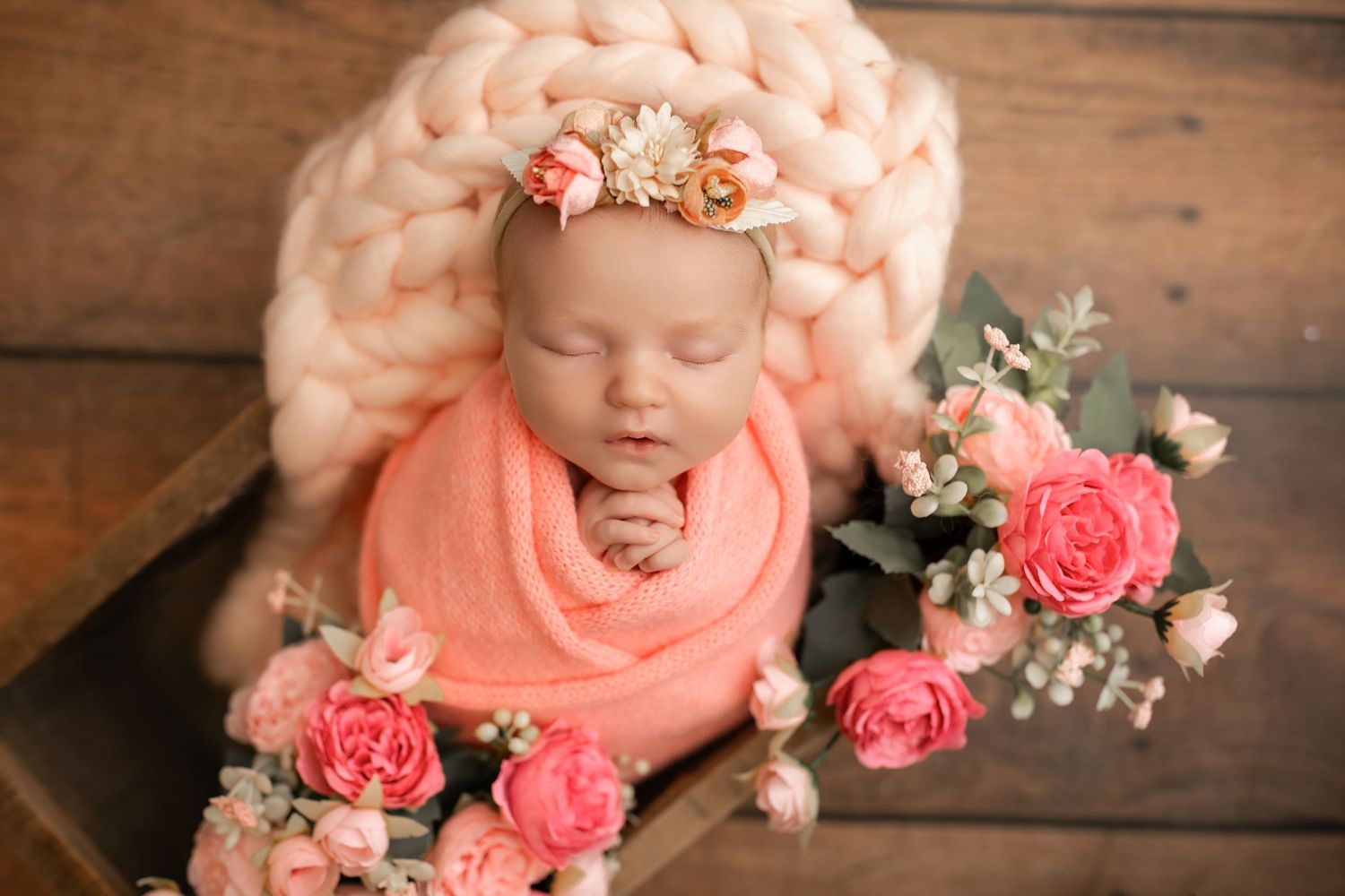 Newborn baby swaddled in peach wrap with floral headband in wooden box surrounded by pink roses.