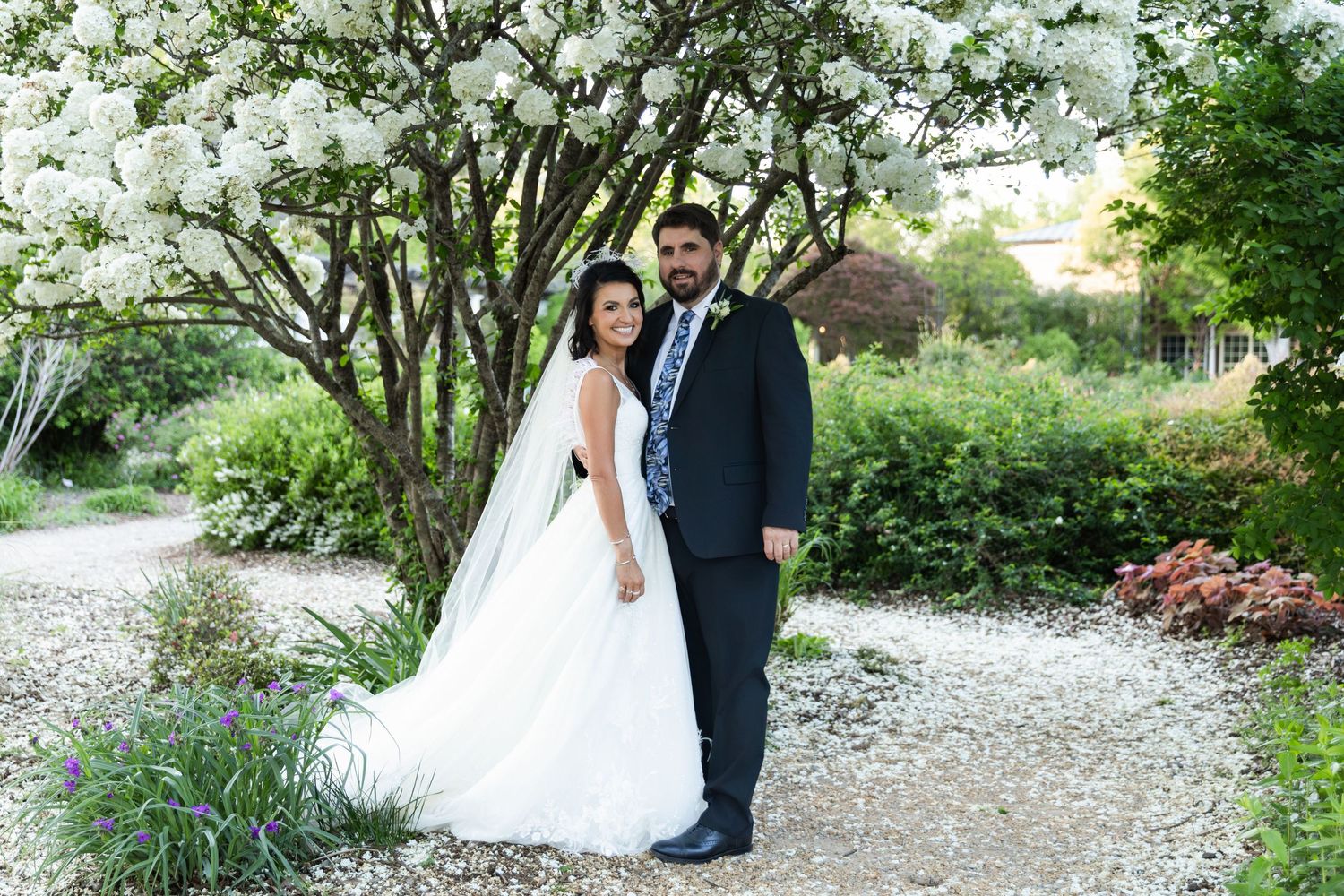 Bride and groom pose together under blooming white tree in lush garden, bride in elegant white gown with veil.