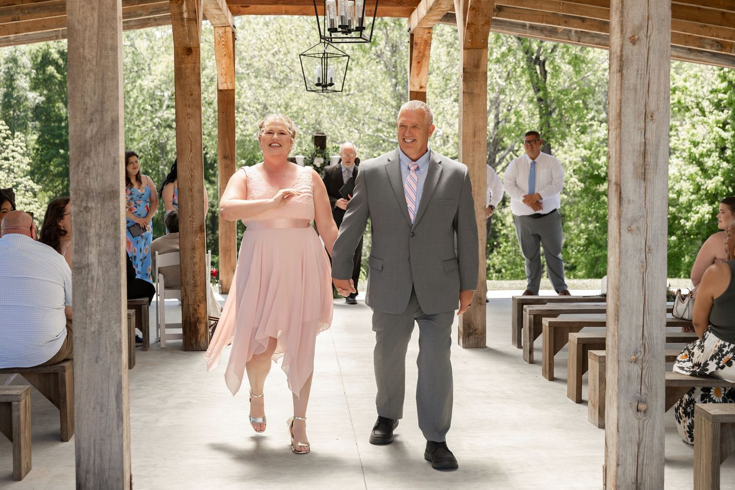Bride and escort walking down the aisle at an outdoor rustic wedding ceremony with wooden benches and lush greenery.