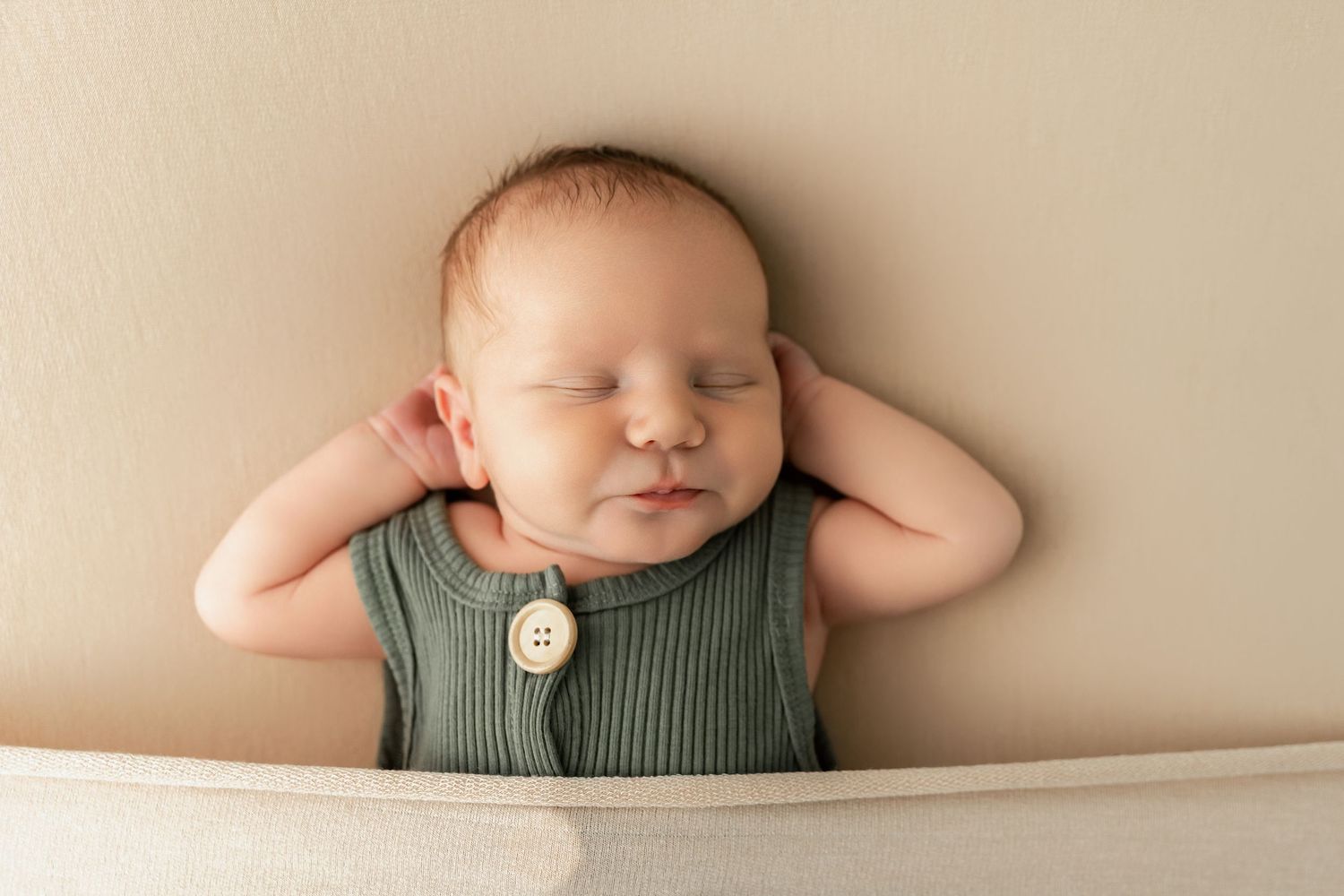 Newborn baby sleeping peacefully in a green ribbed romper with a wooden button, posed with hands behind head.