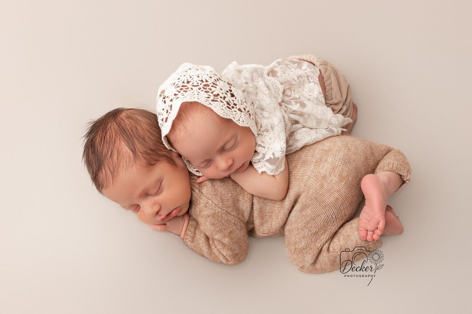 A sleeping baby in a white lace outfit lies on a beige blanket in a professional newborn photography session.