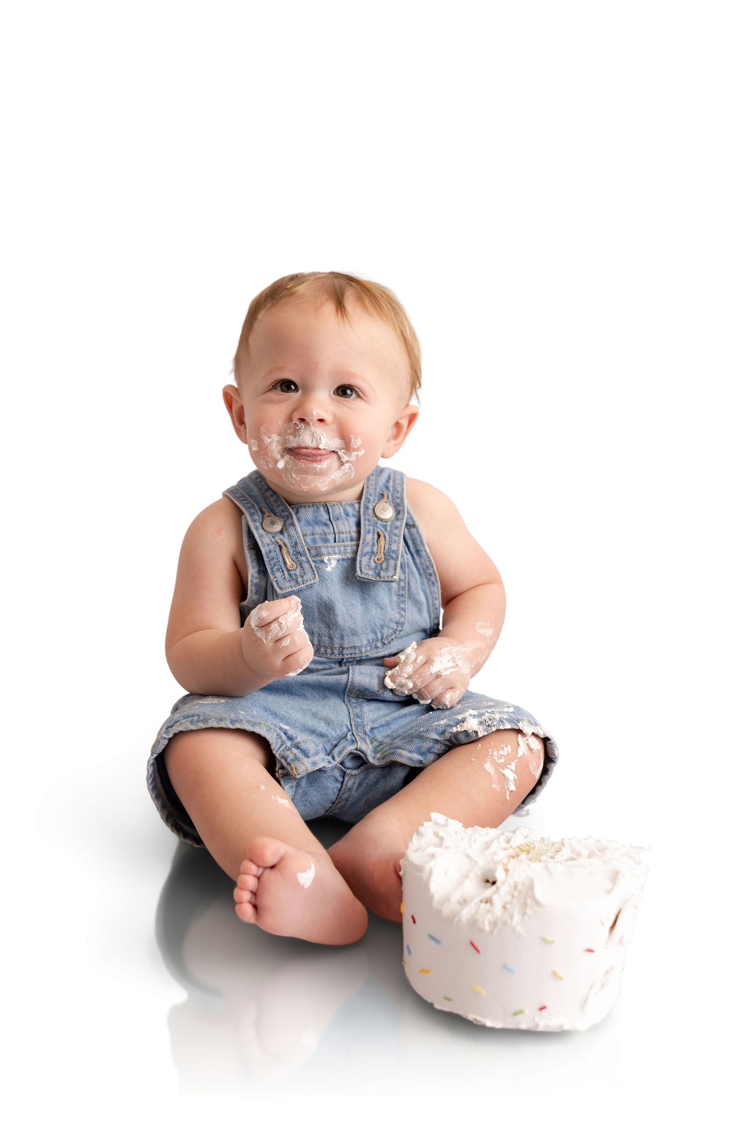 Baby in denim overalls sits barefoot on a white background, smiling with frosting on face and hands beside a small sprinkle smash cake.