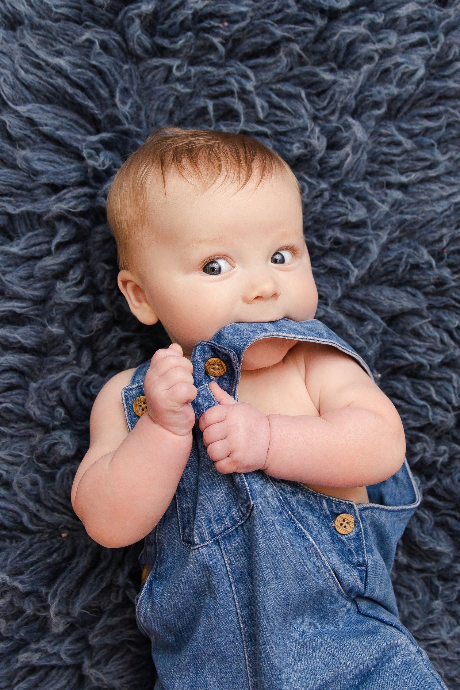 A baby wearing denim overalls lies on a fluffy gray rug while chewing on the straps of their clothing.