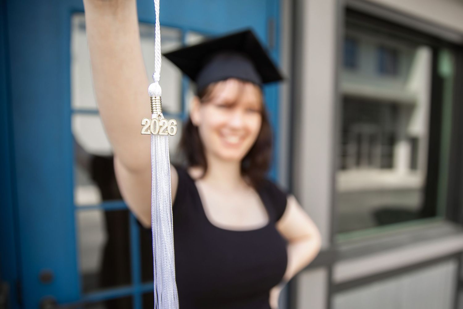 A smiling graduate holds up a 2026 tassel while wearing a black cap and gown outdoors.