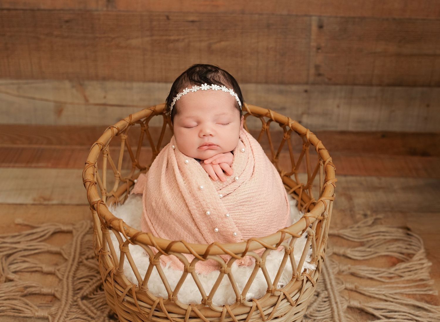 Sleeping newborn baby wrapped in peach swaddle with floral headband posed in wicker basket on rustic wood backdrop.