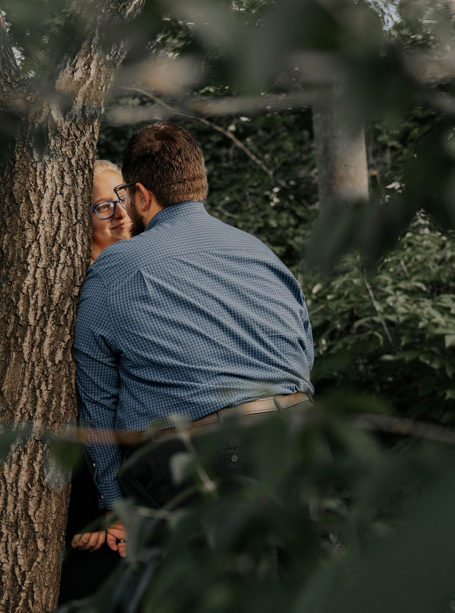 A couple embraces tenderly against a tree trunk in a wooded outdoor setting during golden hour.