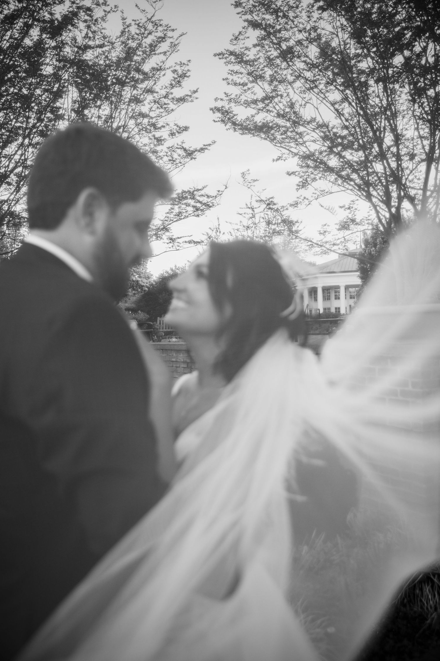 Black and white wedding photo of bride and groom standing close together outdoors, veil flowing, with trees and venue in background.
