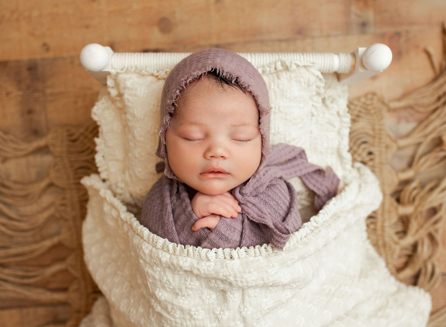 Sleeping newborn baby wrapped in purple swaddle wearing bonnet, resting in white macrame hammock on wood background.