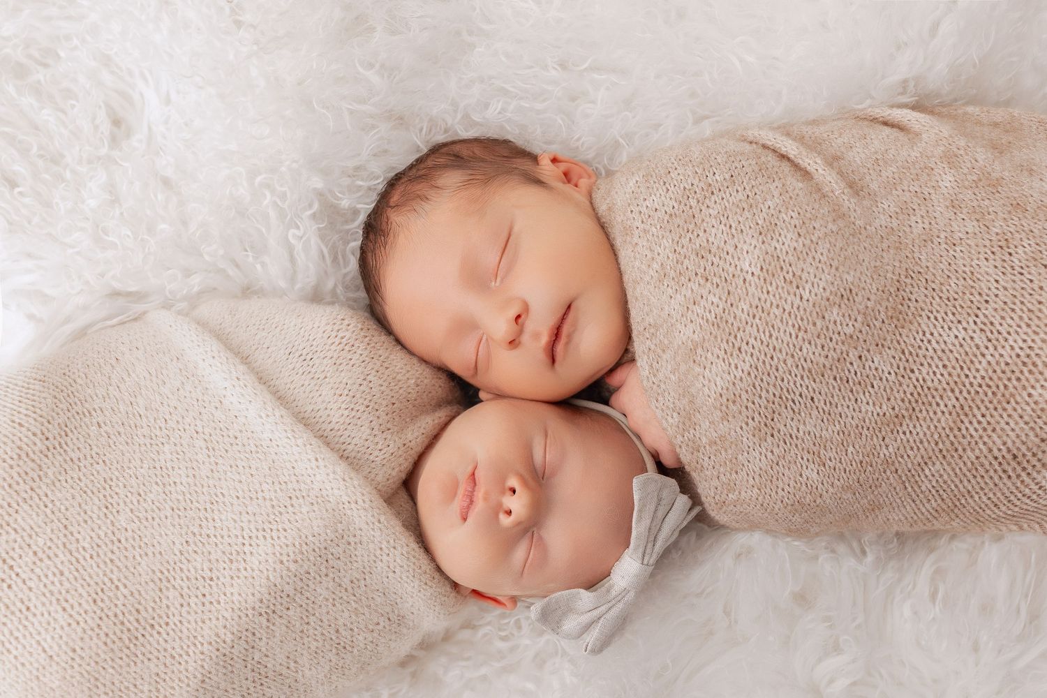 Two sleeping newborn babies wrapped in beige swaddle blankets lie with their heads touching on white fabric.