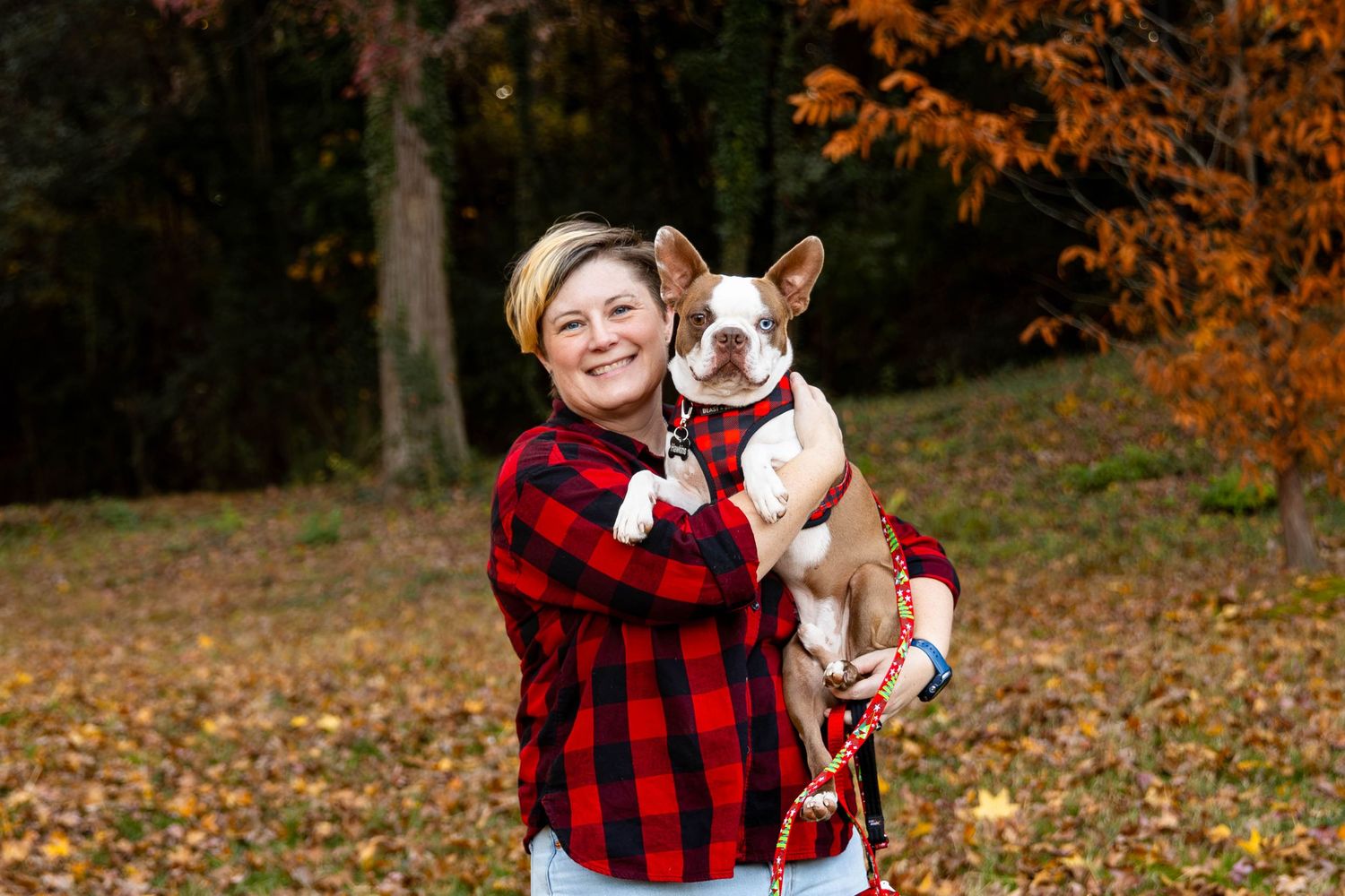A person in a red buffalo plaid flannel shirt poses with a white and brown dog in a fall setting with autumn leaves.