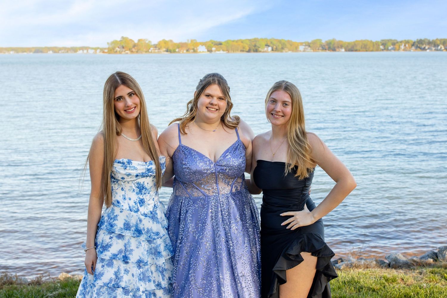 Three young women in formal dresses pose together by a lakeside on a sunny day.