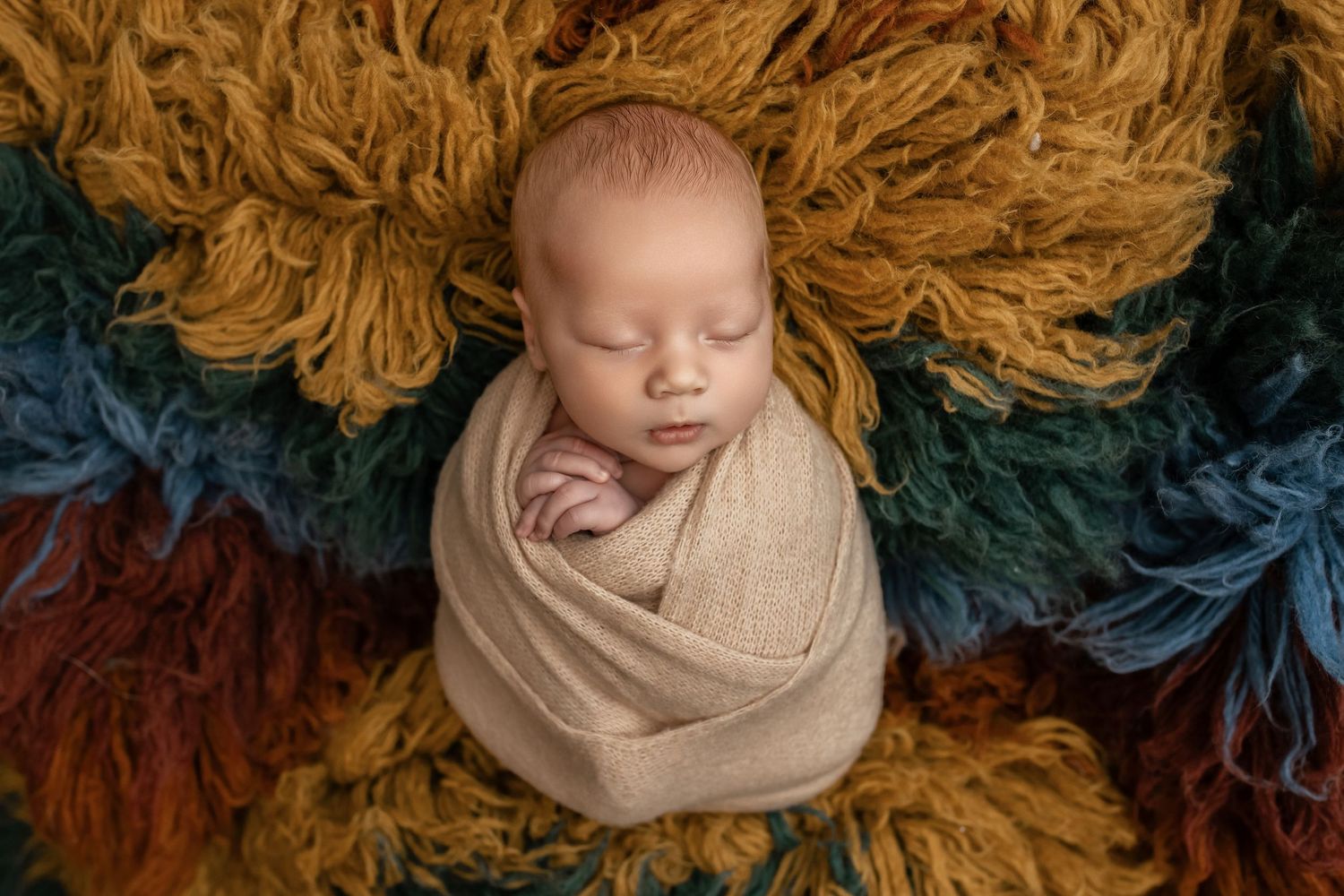 Newborn baby swaddled in beige wrap sleeping peacefully on colorful rainbow flokati rug in professional studio photo.