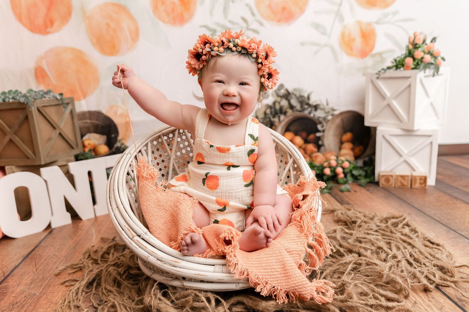 Smiling baby in peach outfit and floral crown sits in wicker basket with orange harvest decor for first birthday photo.