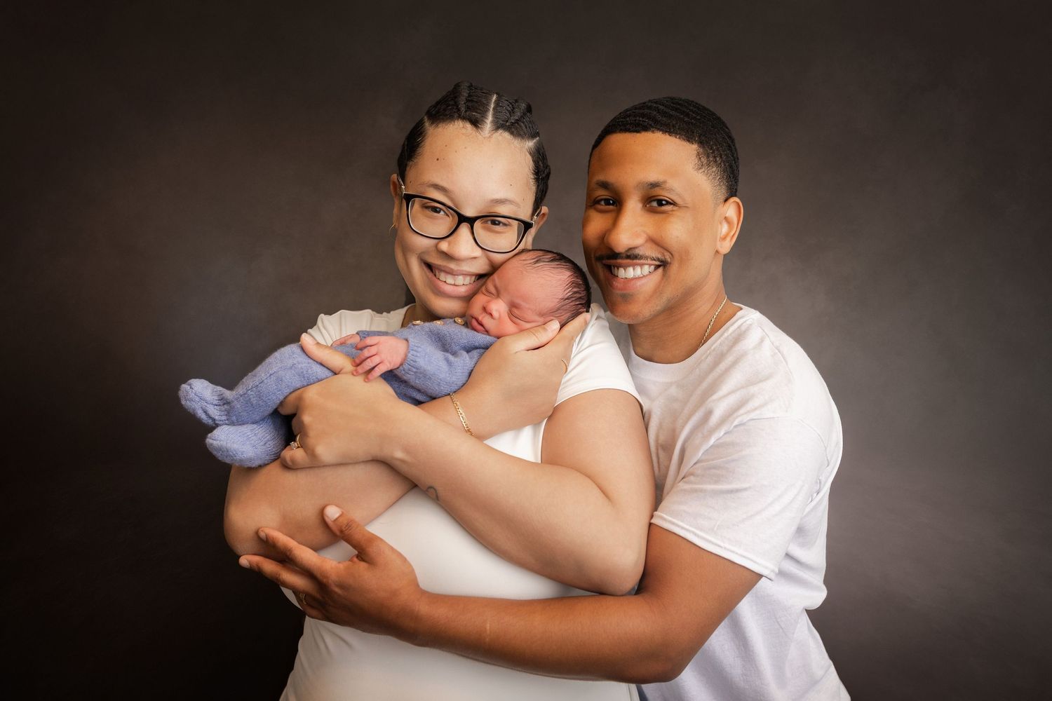 A smiling couple proudly holds their newborn baby in a professional studio portrait photo.