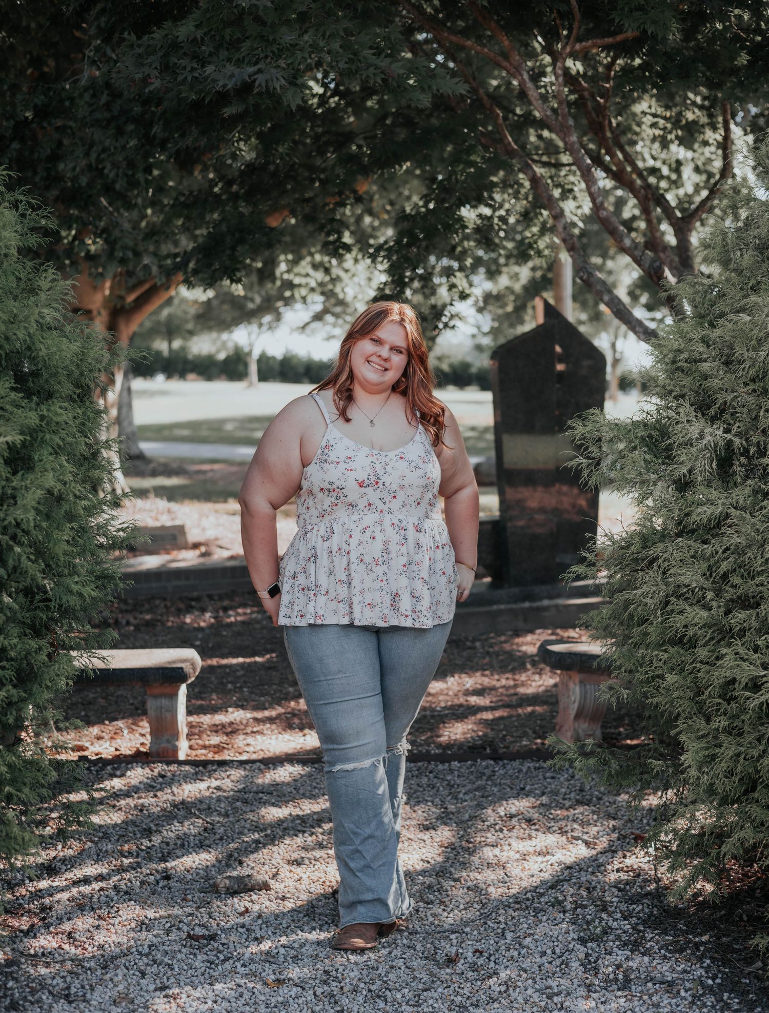 A person wearing a floral top and jeans poses casually in a garden setting with gravel paths and evergreen trees.