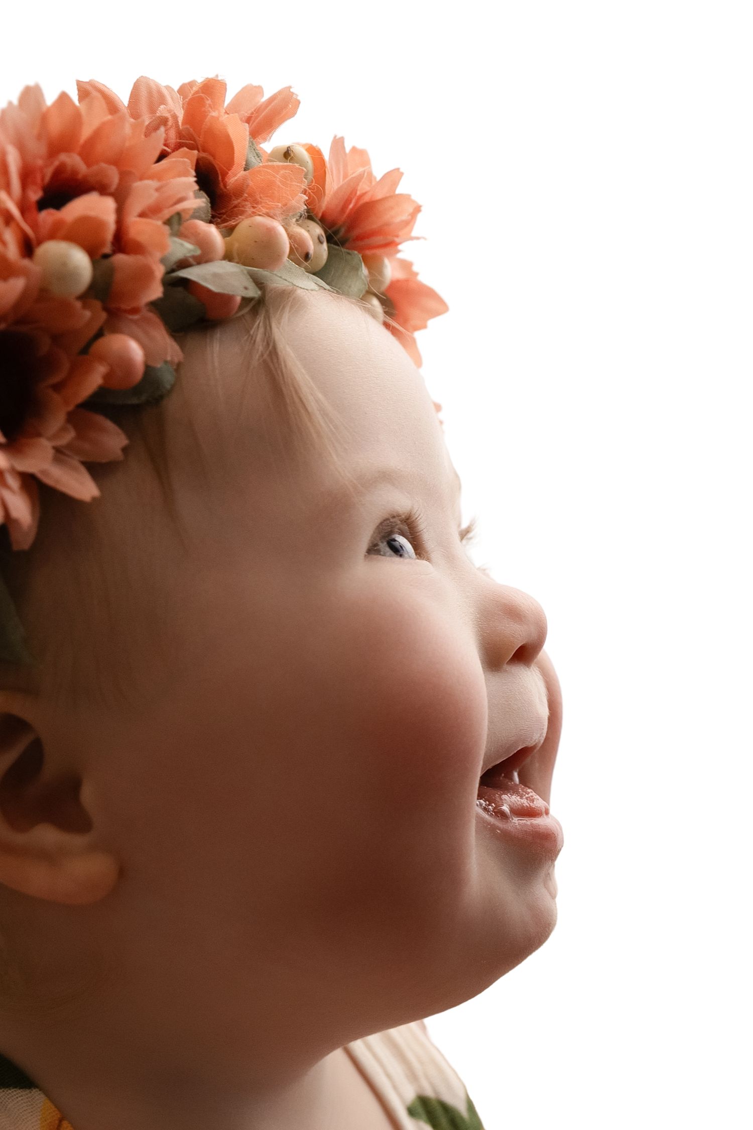 Joyful baby girl wearing a floral crown of orange daisy flowers, laughing and looking up against white background.