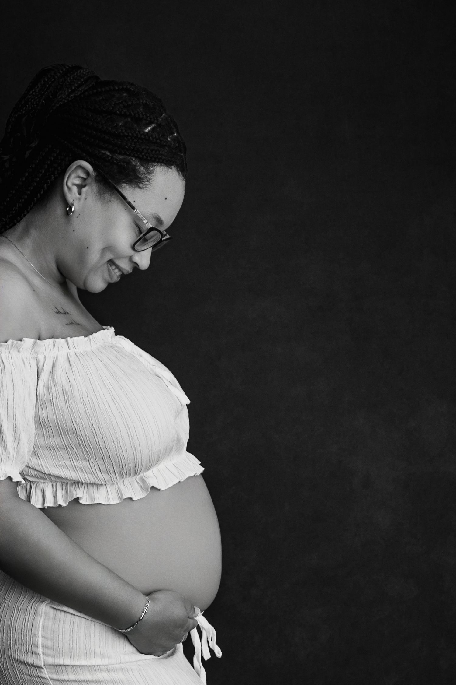 Black and white maternity portrait of a pregnant woman in white off-shoulder dress, cradling her baby bump.