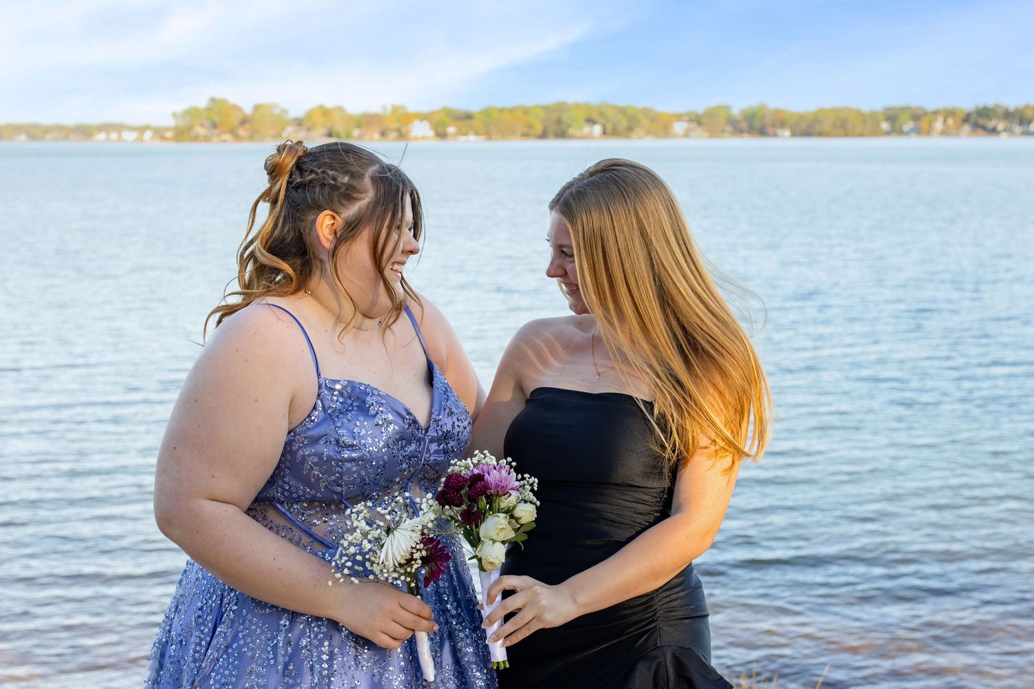 Two young women in formal dresses smile at each other by a lake, one in blue sequins holding a bouquet.