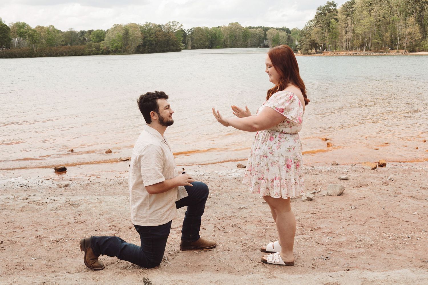 Man kneeling on sandy beach proposing to surprised woman in floral dress near lake surrounded by trees.