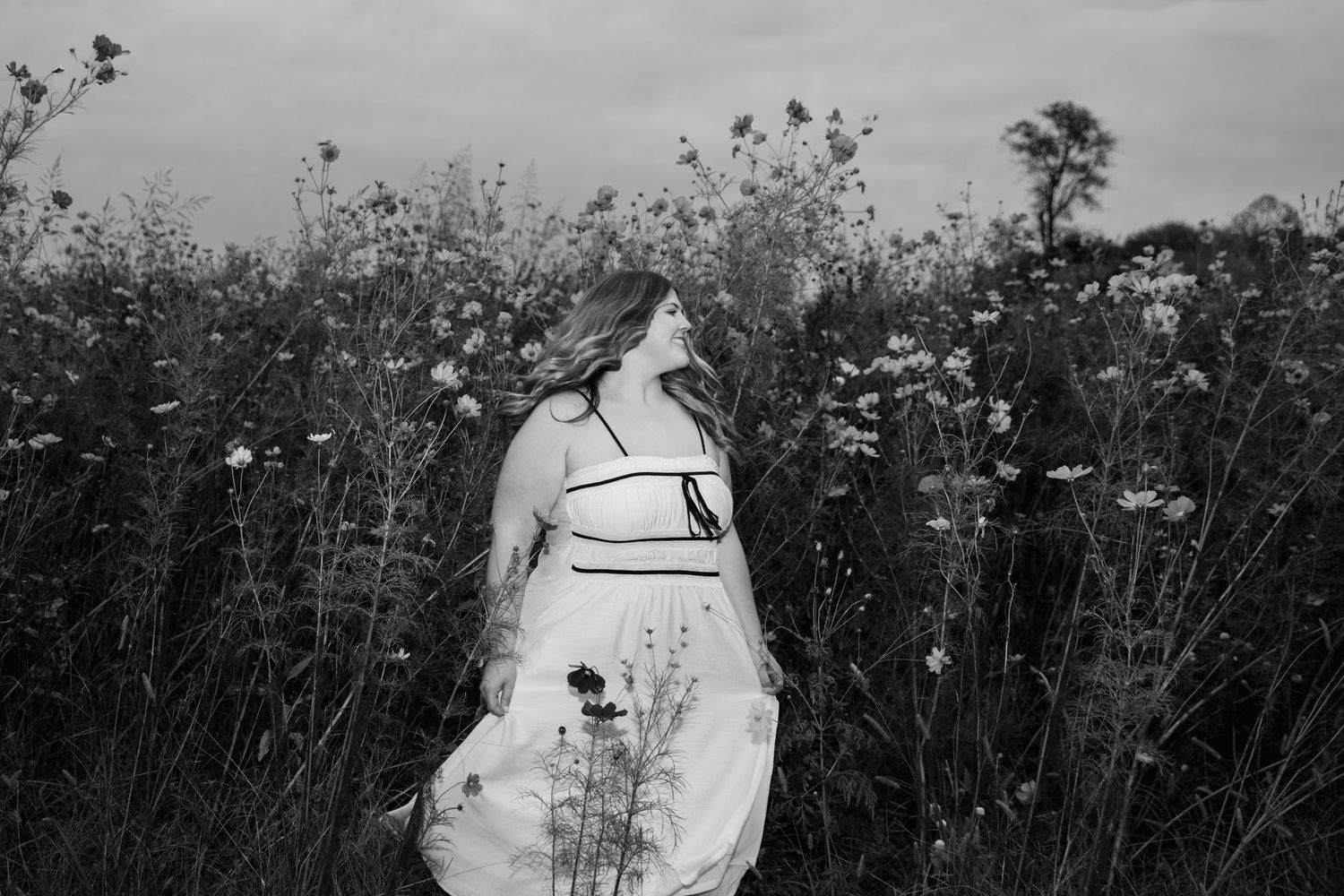 Black and white photo of a woman in a light dress with long hair standing in a tall wildflower meadow, gazing off to the side.