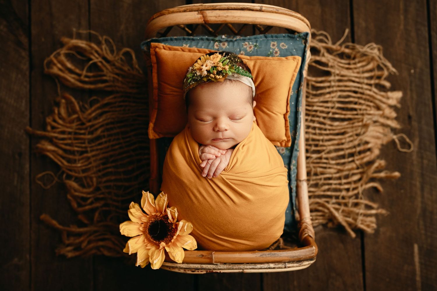 Sleeping newborn baby wrapped in orange swaddle with floral headband, resting in a wooden bed with a sunflower on a rustic floor.