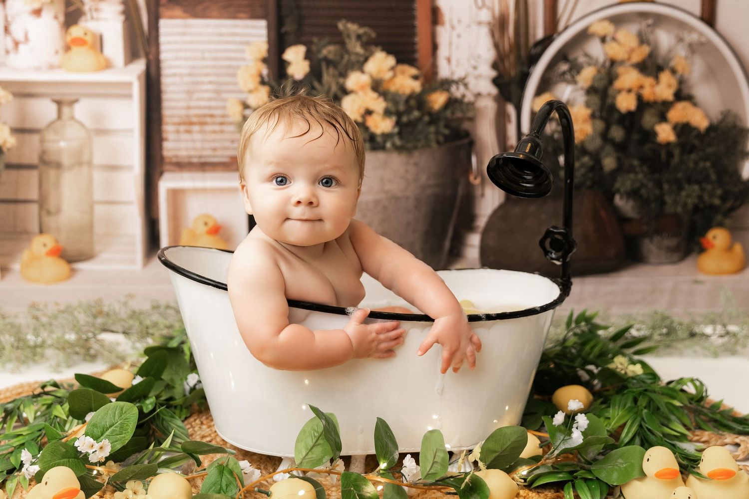 Baby sitting in a vintage white bathtub surrounded by greenery and yellow ducks in a charming photo studio setup.