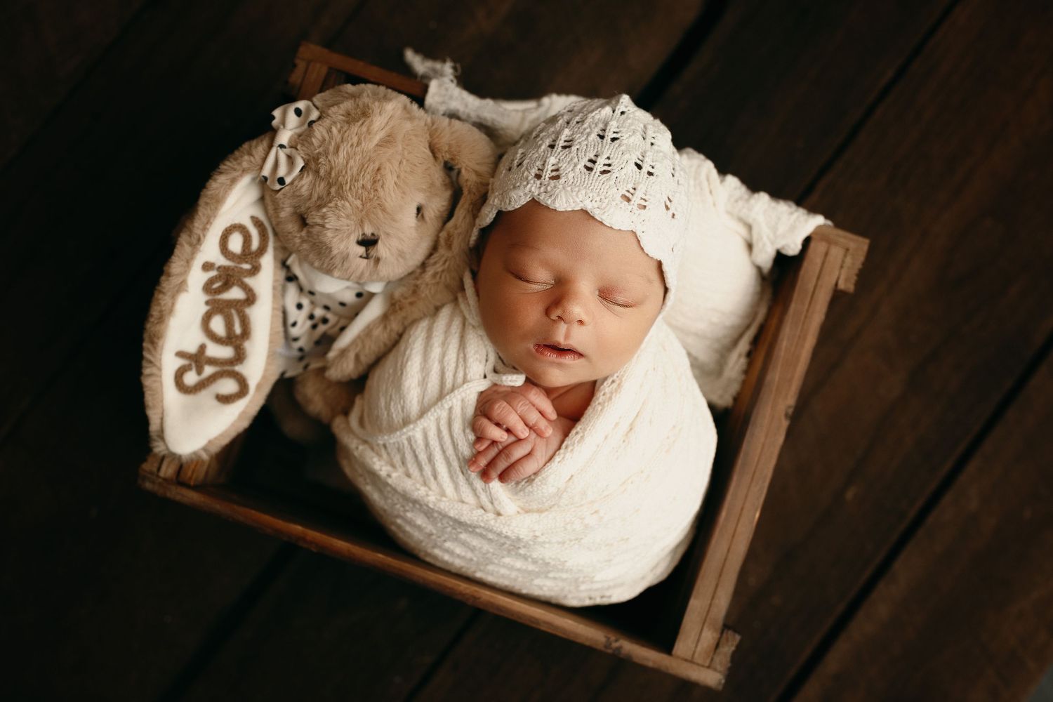 Sleeping newborn baby in a white knit bonnet and cream wrap lies in a wooden crate beside a plush bunny toy with the name Stevie.