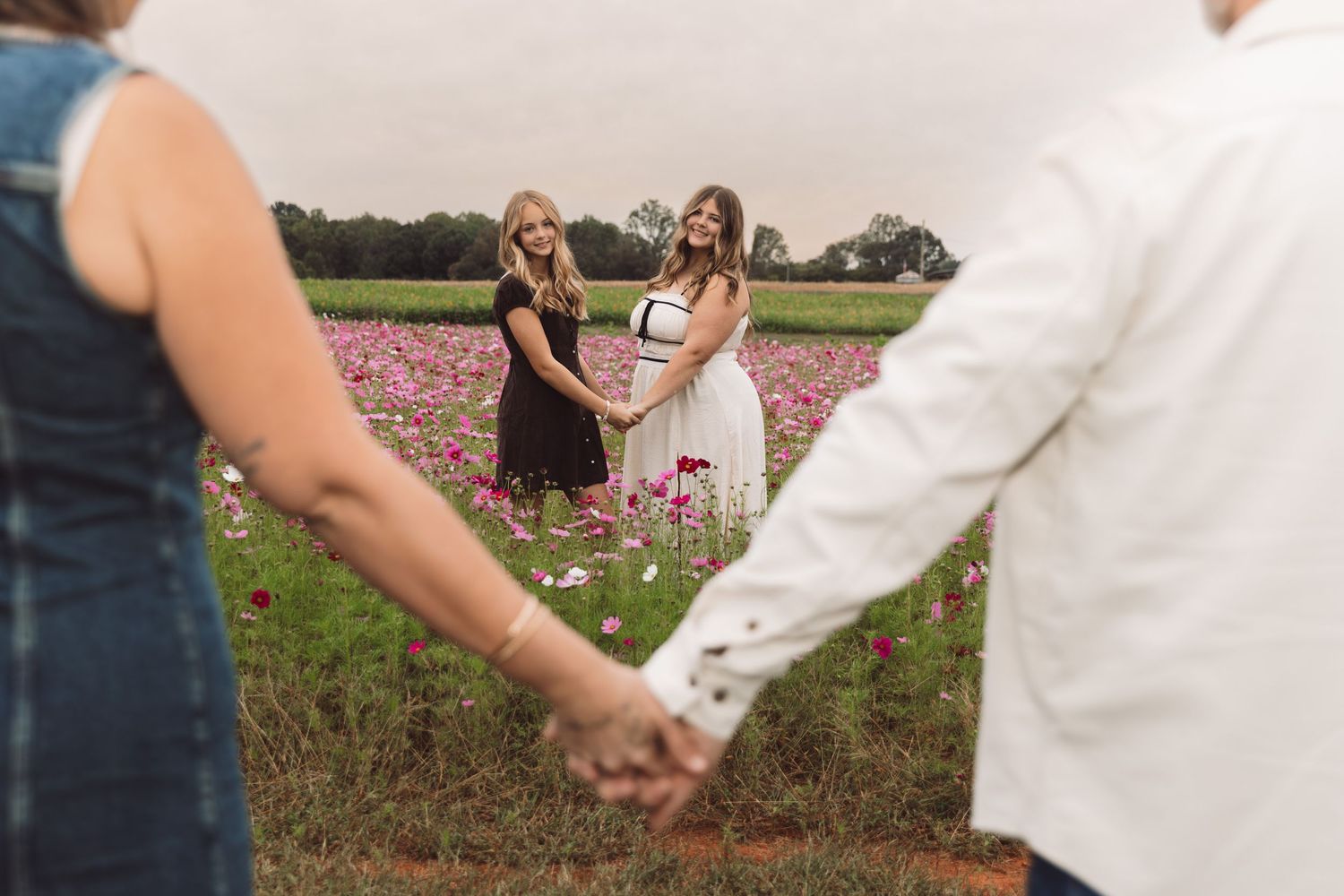 Two women in dresses hold hands in a blooming pink flower field while two people in the foreground join hands to form a circle.